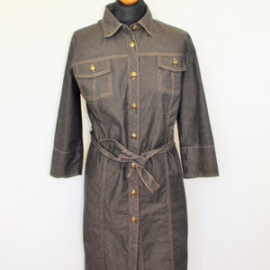 May include: A dark denim shirt dress with a button-up front, long sleeves, and a belt. The dress has two flap pockets on the chest and brown stitching details.