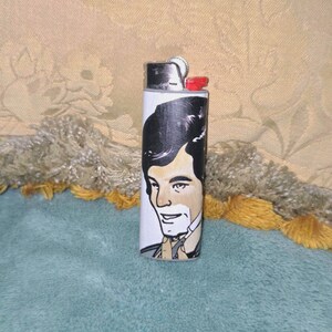 May include: A white lighter featuring a hand-painted portrait of a man with dark hair and a light complexion. The lighter has a silver metal top and a red ignition button. The background is a light yellow fabric.