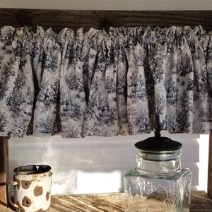 May include: A rustic wooden shelf with a toile-patterned valance. The valance is white with a black pastoral scene. A clear glass jar with a black lid and a distressed metal mug with brown spots sit on the shelf.