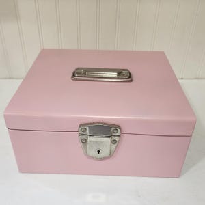 May include: A pink metal box with a rectangular shape. It has a silver handle on top and a silver clasp with a keyhole on the front. The box is sitting on a white surface.