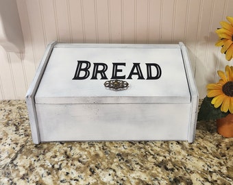 May include: A white wooden bread box with the word "BREAD" in black lettering on the front. The box has a decorative metal clasp and is next to a small pot of sunflowers.