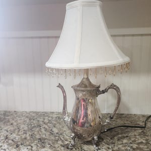 May include: A table lamp featuring a silver-toned metal teapot base with an ornate handle, curved spout, and three decorative feet. The lamp is topped with a white fabric lampshade adorned with a fringe of small, clear, dangling crystal beads.