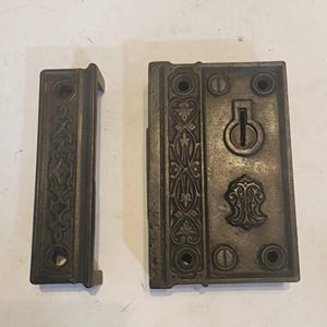 May include: A dark metal door latch and strike plate set featuring intricate embossed floral patterns. The rectangular latch mechanism includes a central keyhole and two screw holes, while the matching strike plate features a narrow vertical design.
