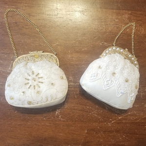 May include: Two vintage ivory-colored purses with gold-tone metal frames and chain handles. One purse features a floral design with beaded accents, while the other has a lace overlay with pearl embellishments.