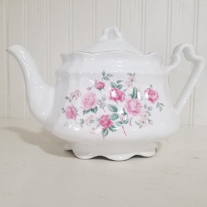 May include: White porcelain teapot with a lid, featuring a floral design of pink roses and green leaves. The teapot has a curved spout and a heart-shaped handle. A decorative piece for serving tea.