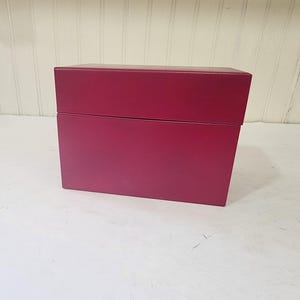 May include: A rectangular, magenta-colored box with a lid. The box is sitting on a white surface. The box is made of a solid material and has a smooth, matte finish. The lid sits flush with the base of the box.