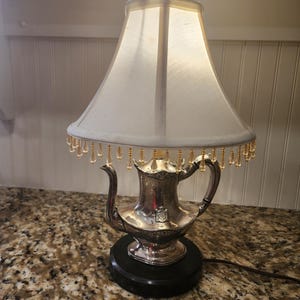 May include: A table lamp featuring a silver-toned metal teapot base with a handle and spout, mounted on a circular black pedestal. The lamp has a white fabric shade adorned with a fringe of small, clear, teardrop-shaped glass beads.