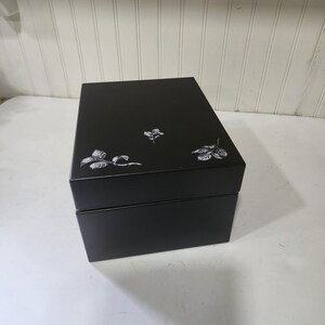 May include: A black box with a hinged lid. The box is decorated with three white floral designs. The box is rectangular and appears to be made of a solid material.