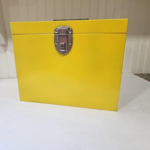 May include: A bright yellow rectangular metal storage box with a smooth finish. The box features a silver-toned metal latch mechanism centered on the front lid, designed to keep the container securely closed. It has a flat top and base.