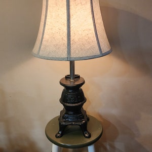 May include: A black cast iron lamp with a light beige lampshade. The lamp base is shaped like a small stove and sits on a green stool with white legs. The lampshade has a light blue trim.