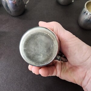 May include: A collection of silver-colored metal cups with ornate handles. One cup is held in a hand, showcasing the bottom. The cups have a reflective surface and a classic design, suggesting they are vintage or antique.
