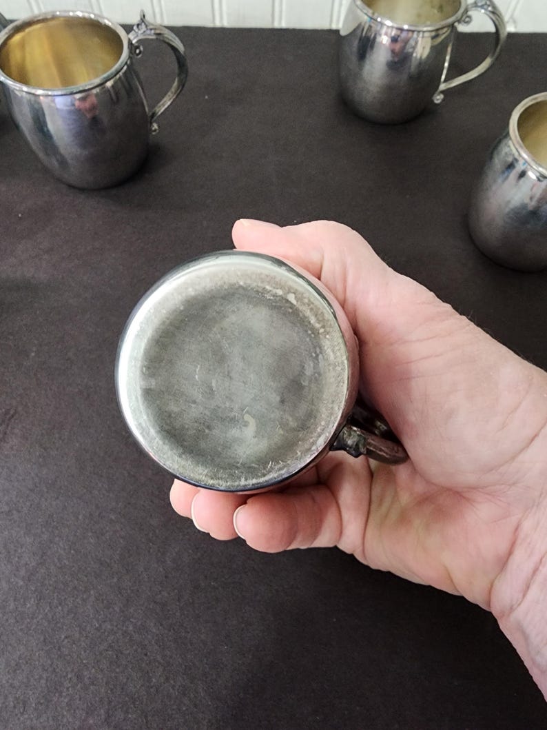 May include: A collection of silver-colored metal cups with ornate handles. One cup is held in a hand, showcasing the bottom. The cups have a reflective surface and a classic design, suggesting they are vintage or antique.