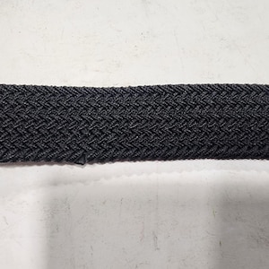 May include: Close-up of a black woven belt. The belt has a textured, herringbone pattern. The material appears to be a durable, elastic fabric. The belt is displayed against a white background.