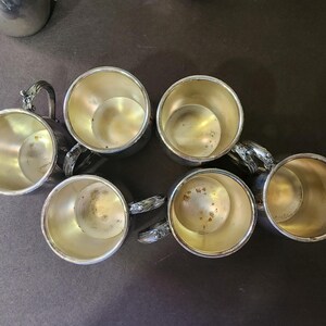 May include: Six silver-colored cups with ornate handles are arranged on a dark surface. The cups have a shiny interior and a detailed handle design. The cups are likely for drinking beverages.