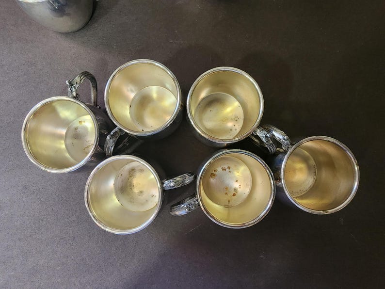 May include: Six silver-colored cups with ornate handles are arranged on a dark surface. The cups have a shiny interior and a detailed handle design. The cups are likely for drinking beverages.