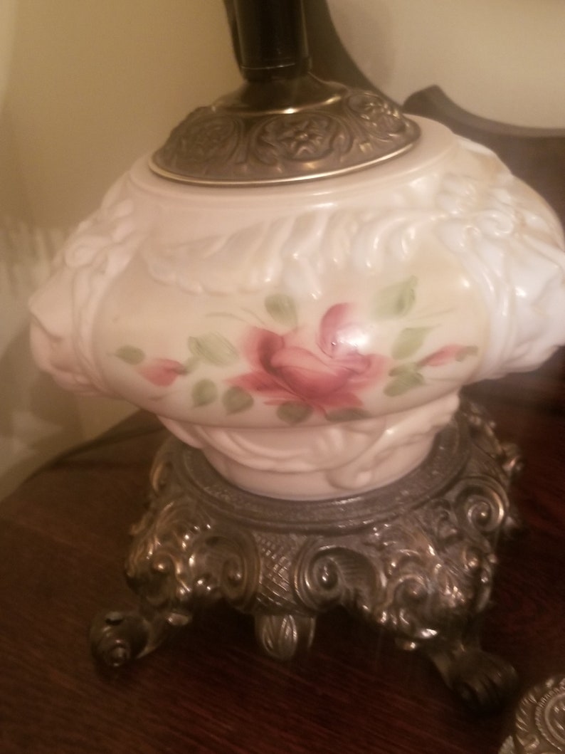 K&ouml;nnte beinhalten: Eine Tischlampe im Vintage-Stil mit einem wei&szlig;en Glasschirm mit Blumenmuster und einem silberfarbenen Metallsockel. Der Schirm zeigt ein handgemaltes Design einer rosa Rose mit gr&uuml;nen Bl&auml;ttern. Der Sockel hat verzierte, gerollte Details.