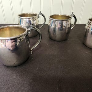 May include: A collection of silver-colored metal mugs with ornate handles. The mugs have a rounded shape with a gold-colored interior. The handles have a decorative floral design. The mugs are arranged on a dark surface.