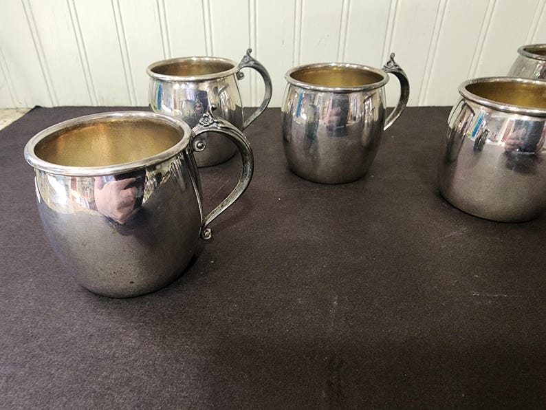 May include: A collection of silver-colored metal mugs with ornate handles. The mugs have a rounded shape with a gold-colored interior. The handles have a decorative floral design. The mugs are arranged on a dark surface.