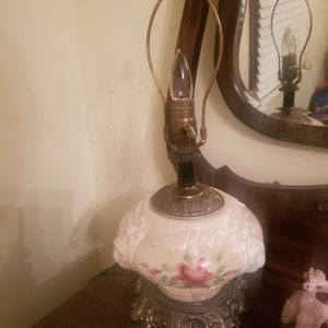 K&ouml;nnte beinhalten: Eine Vintage-Tischlampe mit einem rosa-wei&szlig;en Blumendesign. Die Lampe hat einen dekorativen Sockel und eine Gl&uuml;hbirne mit einem messingfarbenen Rahmen. Die Lampe steht auf einer Holzoberfl&auml;che.
