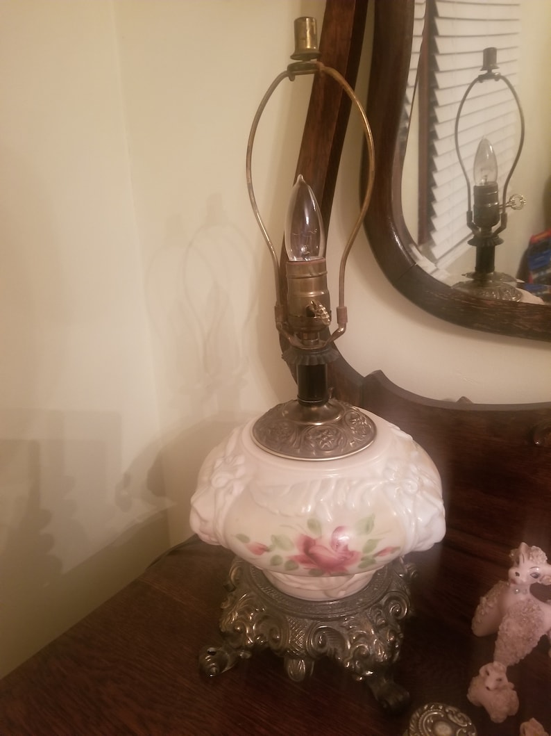 K&ouml;nnte beinhalten: Eine Vintage-Tischlampe mit einem rosa-wei&szlig;en Blumendesign. Die Lampe hat einen dekorativen Sockel und eine Gl&uuml;hbirne mit einem messingfarbenen Rahmen. Die Lampe steht auf einer Holzoberfl&auml;che.