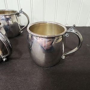 May include: Several silver-colored mugs with ornate handles are arranged on a dark surface. The mugs have a rounded shape and a shiny, reflective surface. The handles feature a decorative floral design.