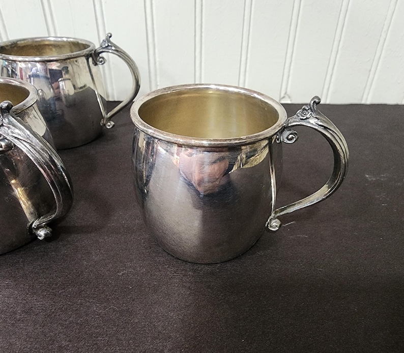 May include: Several silver-colored mugs with ornate handles are arranged on a dark surface. The mugs have a rounded shape and a shiny, reflective surface. The handles feature a decorative floral design.