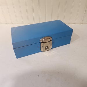 May include: A rectangular blue metal box with a silver clasp and lock. The box is closed and sits on a white surface. The box is approximately 12 inches long.