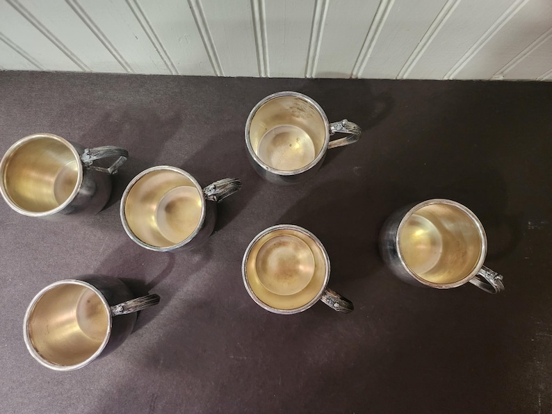 May include: Six silver-colored cups with handles are arranged on a dark surface. The cups have a shiny, metallic exterior and a gold-colored interior. The handles have a decorative design.