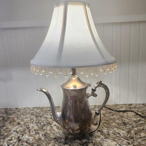 May include: A table lamp constructed from a silver-toned metal teapot base with a curved handle and spout. The lamp features a white, bell-shaped fabric shade adorned with a fringe of clear, teardrop-shaped glass beads hanging from the bottom edge.