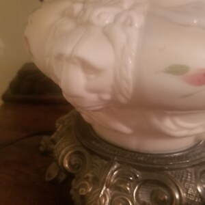 K&ouml;nnte beinhalten: Eine Vintage-Lampe aus wei&szlig;em Glas mit detailliertem Gesichtsdesign und floralen Akzenten. Die Lampe hat einen dekorativen Metallsockel mit aufwendigen Details. Das Design der Lampe erinnert an antike Leuchten.