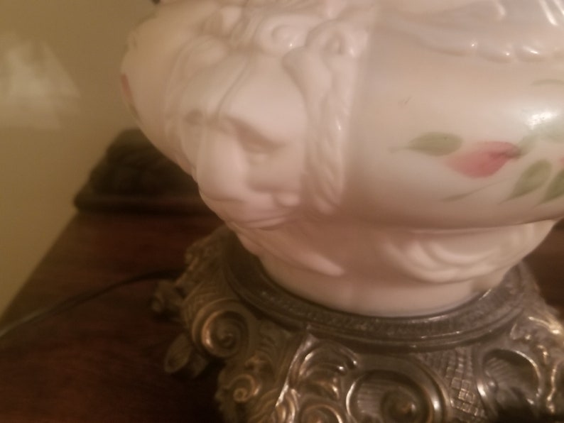 K&ouml;nnte beinhalten: Eine Vintage-Lampe aus wei&szlig;em Glas mit detailliertem Gesichtsdesign und floralen Akzenten. Die Lampe hat einen dekorativen Metallsockel mit aufwendigen Details. Das Design der Lampe erinnert an antike Leuchten.