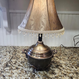 May include: An antique table lamp with a bronze-colored base and a patterned lampshade. The shade is brown with a floral design and a beaded trim. The lamp has a decorative, ornate base with handles and a power cord.