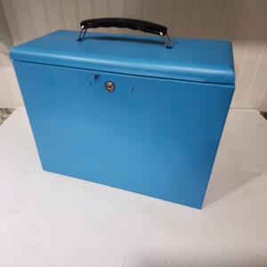 May include: A bright blue, rectangular storage box with a black handle and silver hardware. The box has a lock and keyhole on the front. The box is sitting on a white surface.
