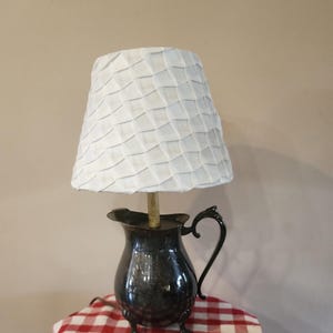 May include: A vintage lamp with a white, textured lampshade. The base is a dark, metallic pitcher with a handle and three small feet. The lamp sits on a red and white checkered tablecloth.
