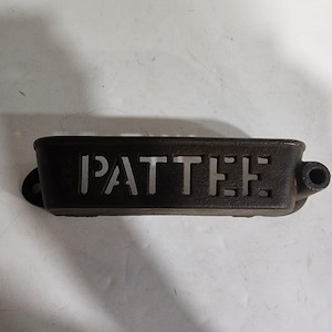 May include: A black cast iron wall-mounted planter with the word "PATTEE" cut out in the center. The planter has a rectangular shape with rounded ends and a small hole on each end for mounting.