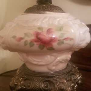 K&ouml;nnte beinhalten: Vintage Tischlampe mit wei&szlig;em Glasschirm mit handgemaltem rosa Rosendesign mit gr&uuml;nen Bl&auml;ttern. Die Lampe hat einen dekorativen, verzierten Metallsockel und einen silberfarbenen Aufsatz.