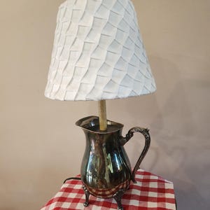 May include: A unique lamp featuring a silver pitcher base with ornate handle and feet. The white lampshade has a textured, geometric design. The lamp sits on a red and white checkered tablecloth.