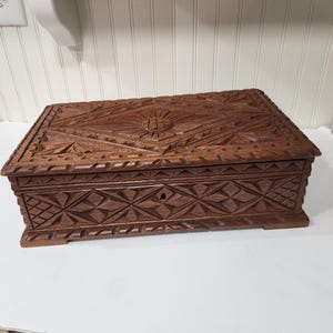May include: A rectangular wooden box with intricate carved geometric and floral patterns. The box is a rich brown color and features a keyhole on the front. The lid is also carved with a sunburst design.