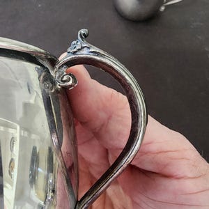 May include: Close-up of a silver creamer with a curved handle and ornate detailing. The creamer has a reflective surface and a small, decorative element at the top of the handle. A smaller silver cup is visible in the background.