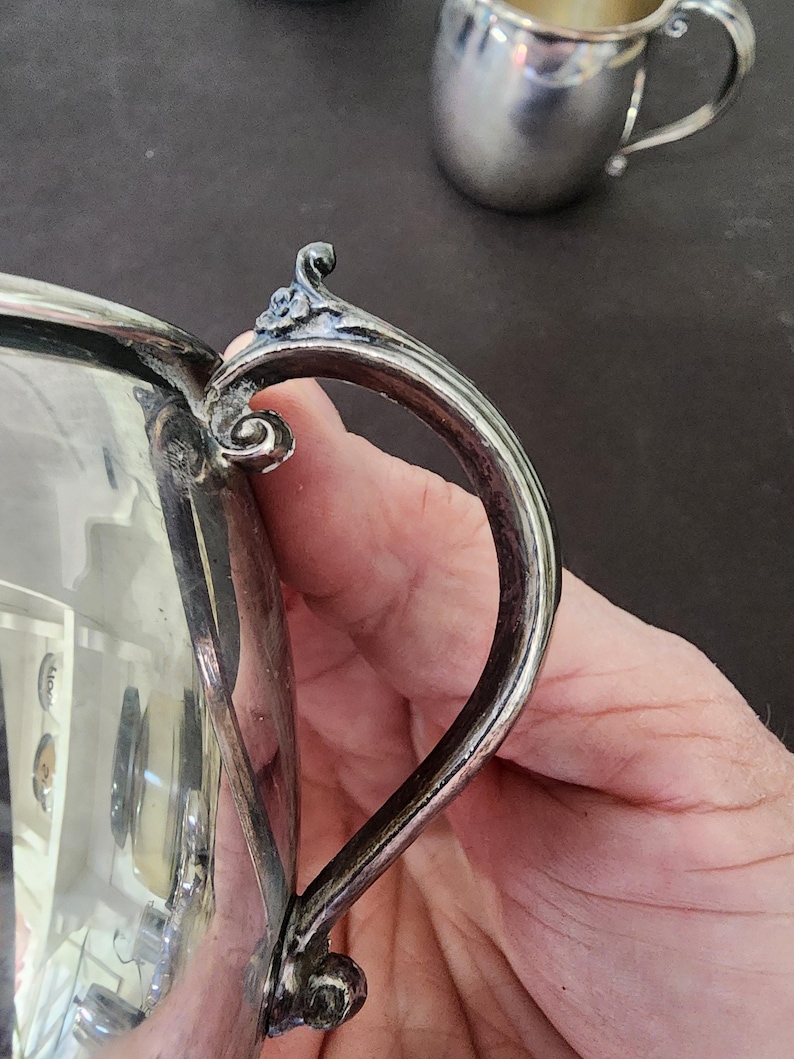 May include: Close-up of a silver creamer with a curved handle and ornate detailing. The creamer has a reflective surface and a small, decorative element at the top of the handle. A smaller silver cup is visible in the background.
