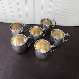 May include: Six silver-colored mugs with small handles are arranged on a dark surface. The mugs have a rounded shape with a shiny interior. The handles have a decorative design. The background is a white paneled wall.