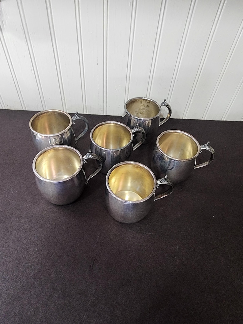 May include: Six silver-colored mugs with small handles are arranged on a dark surface. The mugs have a rounded shape with a shiny interior. The handles have a decorative design. The background is a white paneled wall.