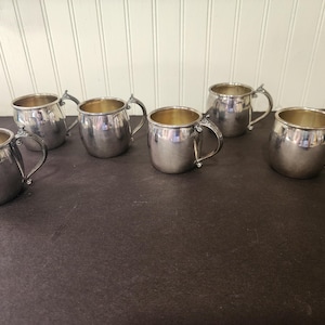 May include: Six silver-colored metal mugs with handles are arranged on a dark surface. The mugs have a rounded shape and a shiny finish. The interior of the mugs appears to be a gold color. The mugs are likely for drinking.