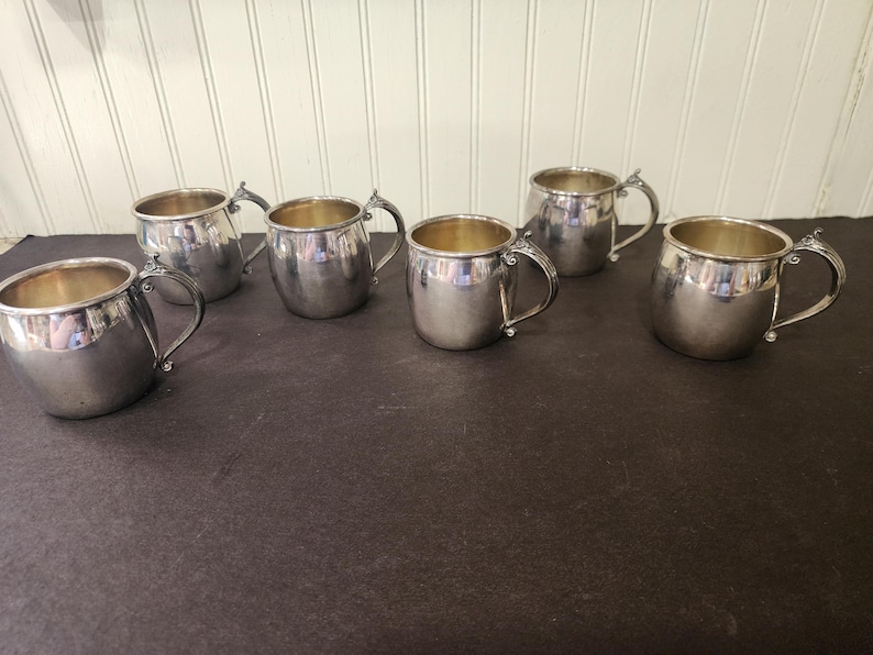 May include: Six silver-colored metal mugs with handles are arranged on a dark surface. The mugs have a rounded shape and a shiny finish. The interior of the mugs appears to be a gold color. The mugs are likely for drinking.