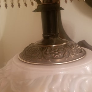 K&ouml;nnte beinhalten: Nahaufnahme einer Vintage-Tischlampe. Die Lampe hat einen wei&szlig;en Glasschirm mit floralem Muster, einen dunklen Metallhals und einen dekorativen, verzierten Sockel. Die Lampe ist mit einem Perlenfransen versehen.