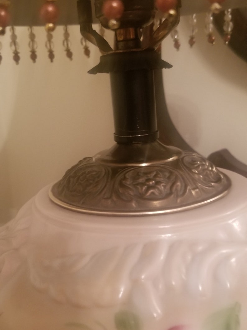 K&ouml;nnte beinhalten: Nahaufnahme einer Vintage-Tischlampe. Die Lampe hat einen wei&szlig;en Glasschirm mit floralem Muster, einen dunklen Metallhals und einen dekorativen, verzierten Sockel. Die Lampe ist mit einem Perlenfransen versehen.