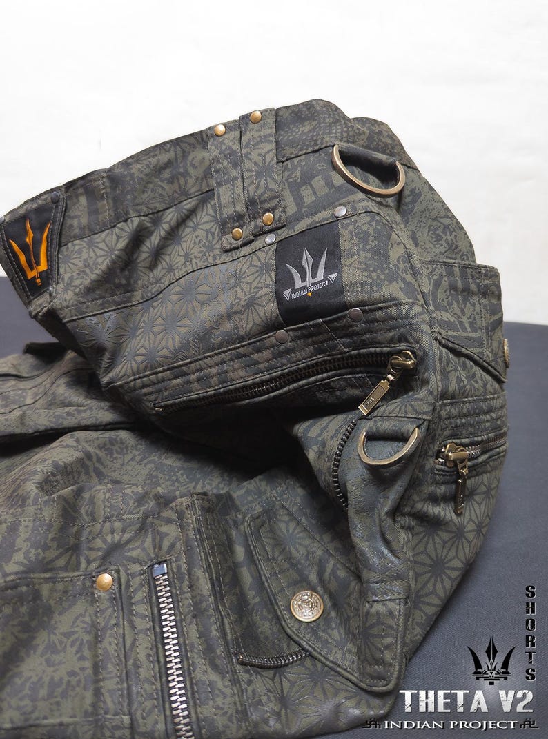 May include: Olive green cargo shorts featuring a repeating geometric star pattern, multiple zippered pockets, brass-toned hardware, and D-ring attachments. A black fabric label displays the text INDIAN PROJECT with a trident logo.