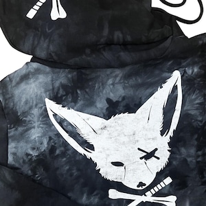 May include: A black and gray tie-dye hoodie featuring a large white graphic of a fox head with an 'X' over one eye, positioned above crossed bones. The hood displays a smaller graphic of crossed bones. The text 'plazma lab.' appears in the corner.