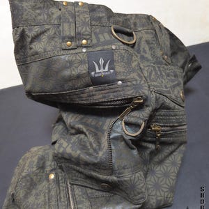 May include: Olive green cargo shorts featuring a dark geometric pattern, multiple zippered pockets, brass-toned hardware, and D-ring attachments. A black fabric label displays the text "INDIAN PROJECT" with a trident logo. The text "THETA V2" is visible.