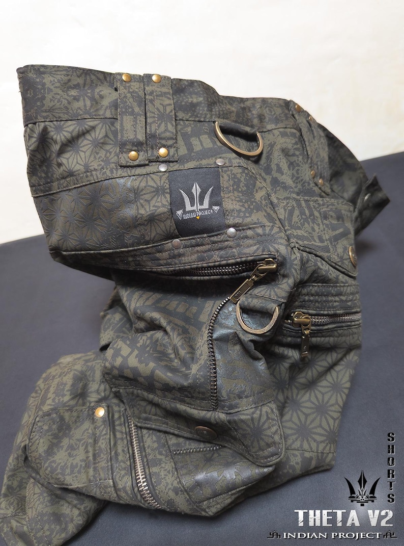May include: Olive green cargo shorts featuring a dark geometric pattern, multiple zippered pockets, brass-toned hardware, and D-ring attachments. A black fabric label displays the text "INDIAN PROJECT" with a trident logo. The text "THETA V2" is visible.